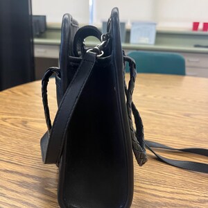 May include: Black leather shoulder bag with a rectangular shape. The bag features a braided handle and a long, adjustable strap. The hardware is silver-toned. The bag is sitting on a wooden table.