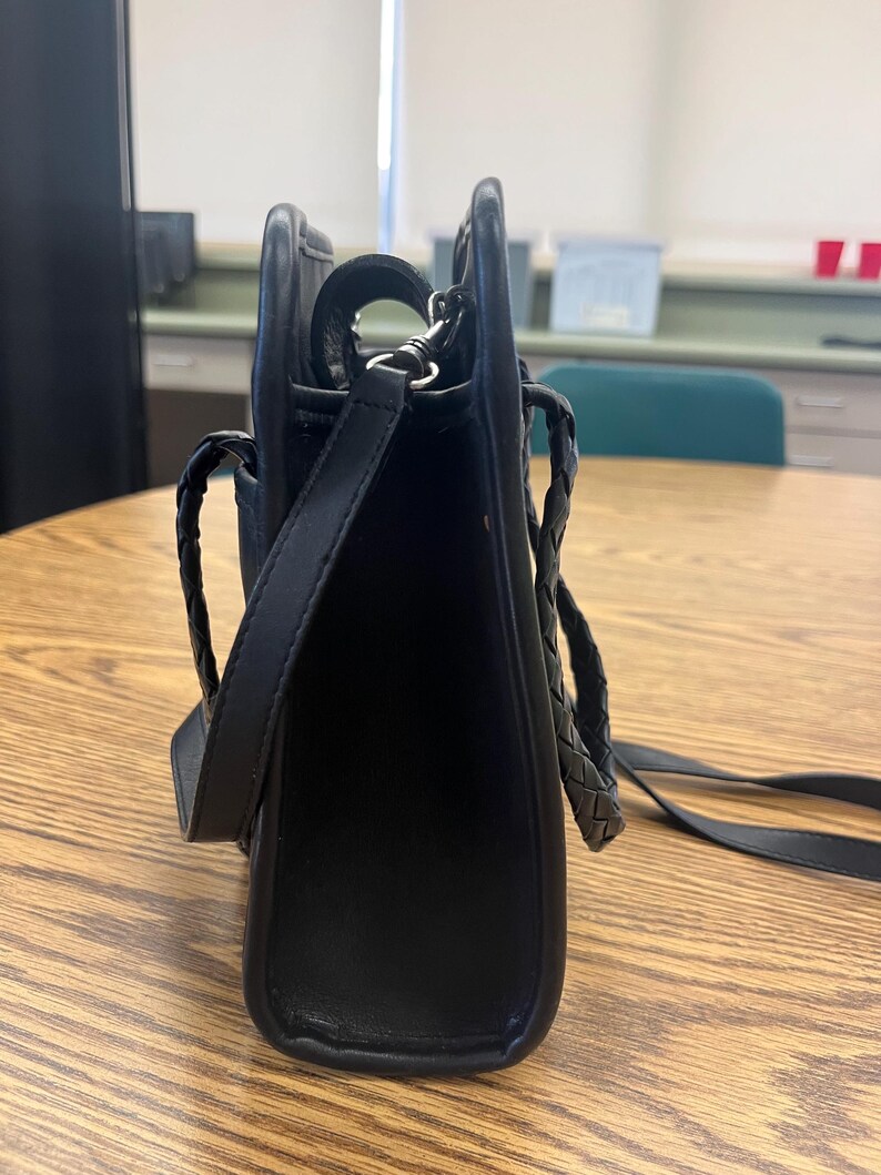 May include: Black leather shoulder bag with a rectangular shape. The bag features a braided handle and a long, adjustable strap. The hardware is silver-toned. The bag is sitting on a wooden table.