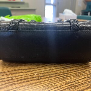 May include: Black handbag with a textured, crocodile-like pattern on the top section. The bag has braided handles and a smooth, solid black body. The bag is rectangular and sits on a wooden surface.