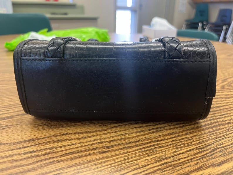 May include: Black handbag with a textured, crocodile-like pattern on the top section. The bag has braided handles and a smooth, solid black body. The bag is rectangular and sits on a wooden surface.