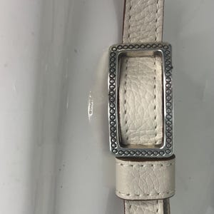 Pode incluir: Close-up de uma correia de couro creme com uma fivela retangular prateada. A correia tem uma superf&iacute;cie texturizada e &eacute; costurada com uma linha contrastante. A fivela &eacute; adornada com pequenos enfeites circulares.