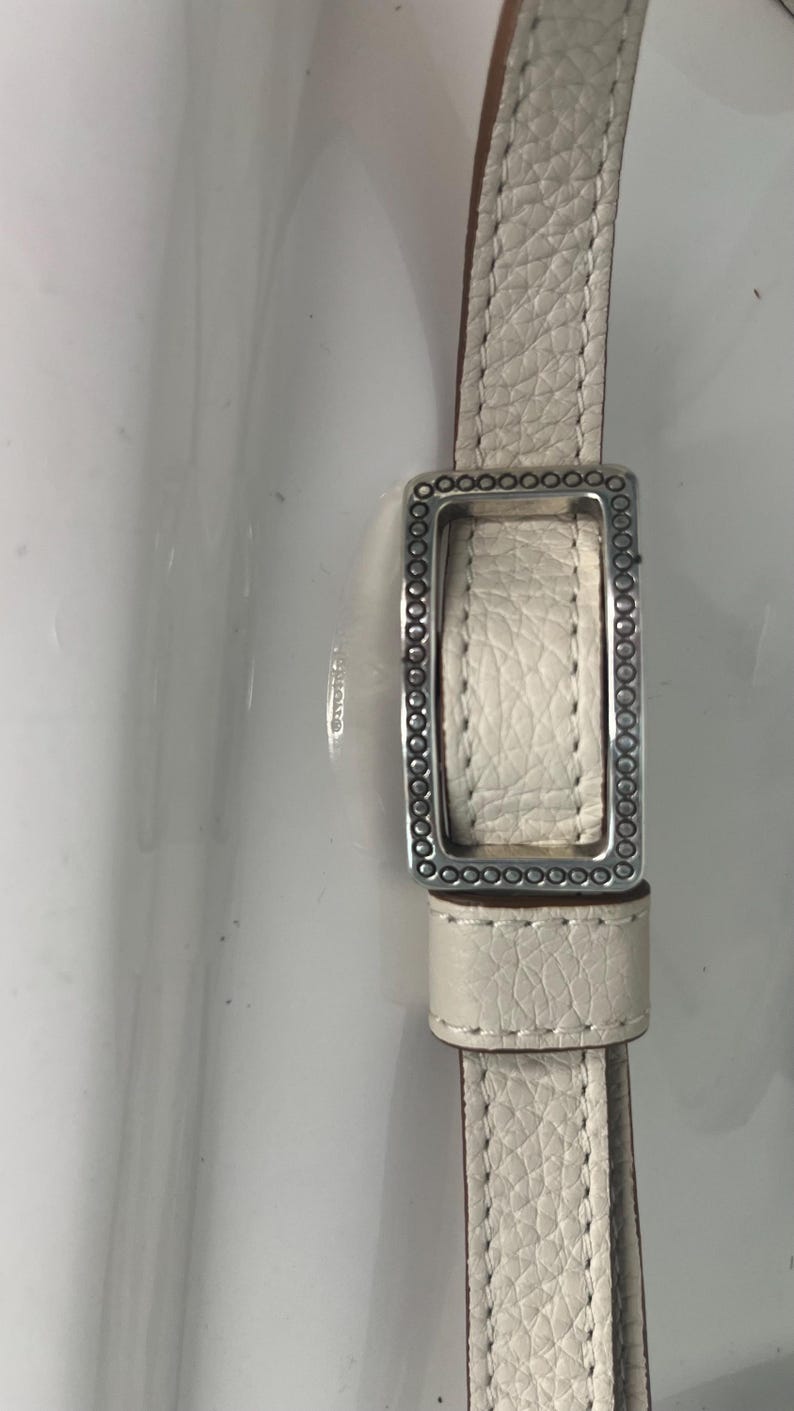 Pode incluir: Close-up de uma correia de couro creme com uma fivela retangular prateada. A correia tem uma superf&iacute;cie texturizada e &eacute; costurada com uma linha contrastante. A fivela &eacute; adornada com pequenos enfeites circulares.