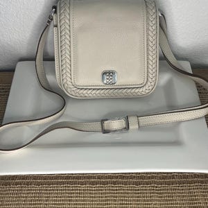Pode incluir: Bolsa transversal creme com uma borda tran&ccedil;ada e al&ccedil;a ajust&aacute;vel. A bolsa apresenta uma fivela prateada e um emblema decorativo na frente. A bolsa &eacute; feita de couro e est&aacute; sobre um prato branco.