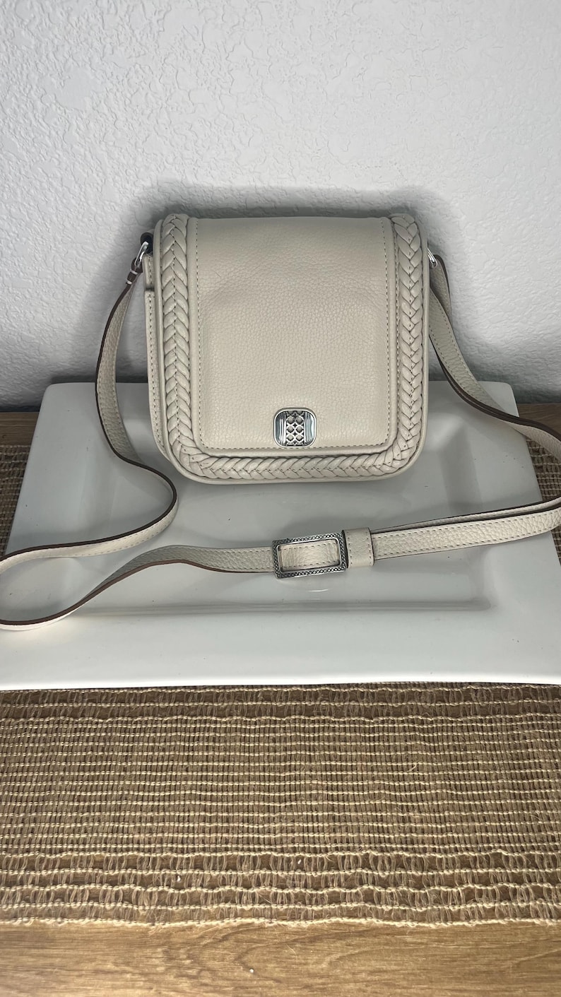 Pode incluir: Bolsa transversal creme com uma borda tran&ccedil;ada e al&ccedil;a ajust&aacute;vel. A bolsa apresenta uma fivela prateada e um emblema decorativo na frente. A bolsa &eacute; feita de couro e est&aacute; sobre um prato branco.