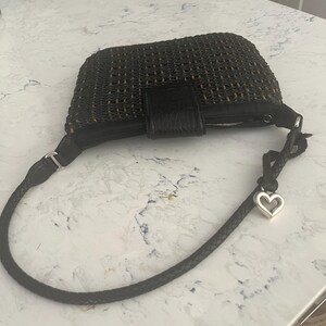 Puede incluir: Bolso de mano tejido negro con cierre de solapa de cuero y correa trenzada. La correa tiene un dije de coraz&oacute;n plateado. El bolso es rectangular y parece hecho de fibras naturales.