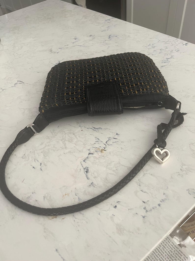 Puede incluir: Bolso de mano tejido negro con cierre de solapa de cuero y correa trenzada. La correa tiene un dije de coraz&oacute;n plateado. El bolso es rectangular y parece hecho de fibras naturales.