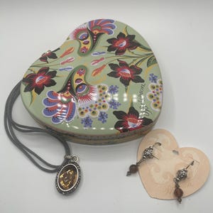 May include: A heart-shaped, floral-patterned trinket box with a matching pendant necklace and earrings. The box is light green with red and purple flowers. The necklace has a brown oval pendant. The earrings have brown beads.