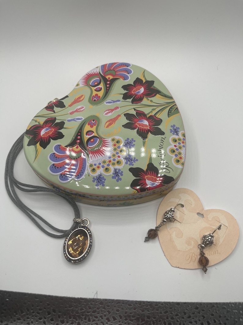 May include: A heart-shaped, floral-patterned trinket box with a matching pendant necklace and earrings. The box is light green with red and purple flowers. The necklace has a brown oval pendant. The earrings have brown beads.