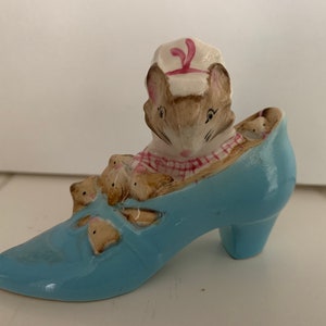 May include: A ceramic figurine of a mouse family in a light blue high-heeled shoe. The mother mouse wears a white chef's hat with a pink accent and a red and white checkered scarf. Several baby mice peek out of the shoe.