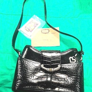 May include: Black handbag with a crocodile-like texture, a silver-tone clasp, and a shoulder strap. The bag has a flap closure and a small silver heart charm. Includes a small card and a tag.