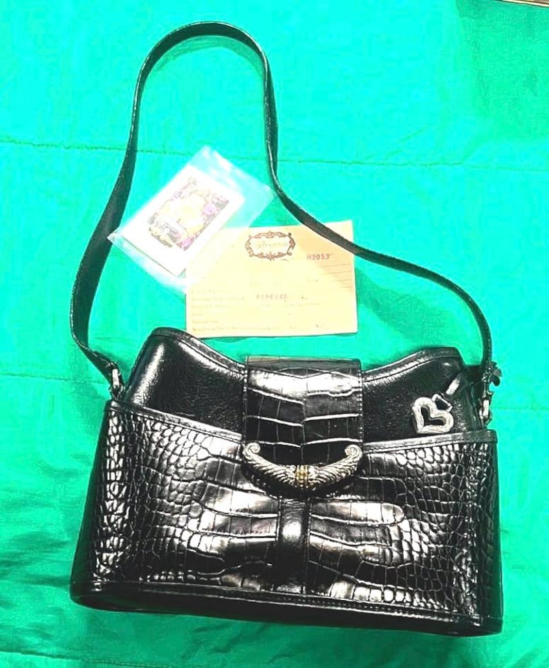 May include: Black handbag with a crocodile-like texture, a silver-tone clasp, and a shoulder strap. The bag has a flap closure and a small silver heart charm. Includes a small card and a tag.