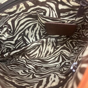 May include: The interior of a handbag featuring a brown and white zebra stripe pattern lining. It includes a zippered top closure, an open slip pocket, and a brown leather patch embossed with the Brighton brand name and descriptive text.