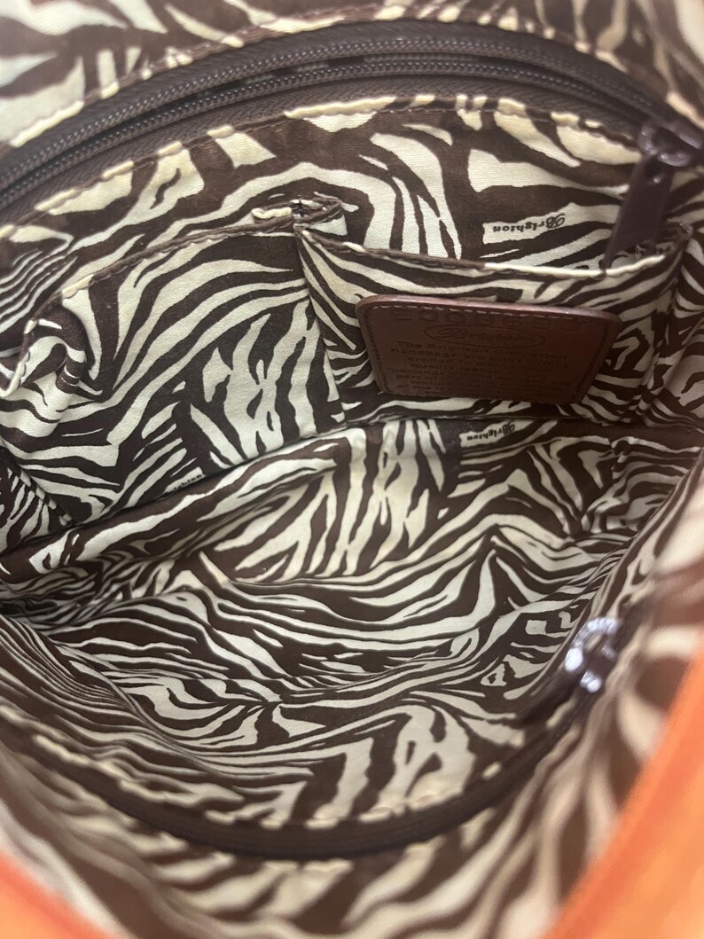 May include: The interior of a handbag featuring a brown and white zebra stripe pattern lining. It includes a zippered top closure, an open slip pocket, and a brown leather patch embossed with the Brighton brand name and descriptive text.