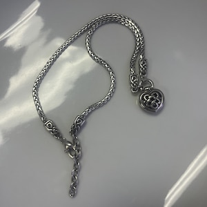 Puede incluir: Collar plateado con un colgante en forma de coraz&oacute;n. El collar presenta un dise&ntilde;o de cadena tejida y un dije decorativo en forma de coraz&oacute;n con un patr&oacute;n calado. El cierre es visible al final de la cadena.