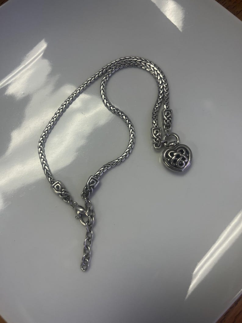 Puede incluir: Collar plateado con un colgante en forma de coraz&oacute;n. El collar presenta un dise&ntilde;o de cadena tejida y un dije decorativo en forma de coraz&oacute;n con un patr&oacute;n calado. El cierre es visible al final de la cadena.