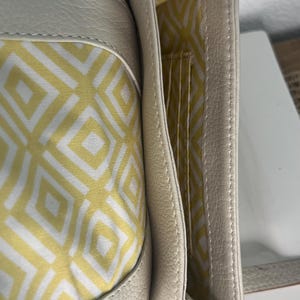 Pode incluir: Bolsa crossbody de couro creme com interior estampado geom&eacute;trico amarelo e branco. A bolsa possui fecho de aba, al&ccedil;a de ombro e v&aacute;rios compartimentos internos.
