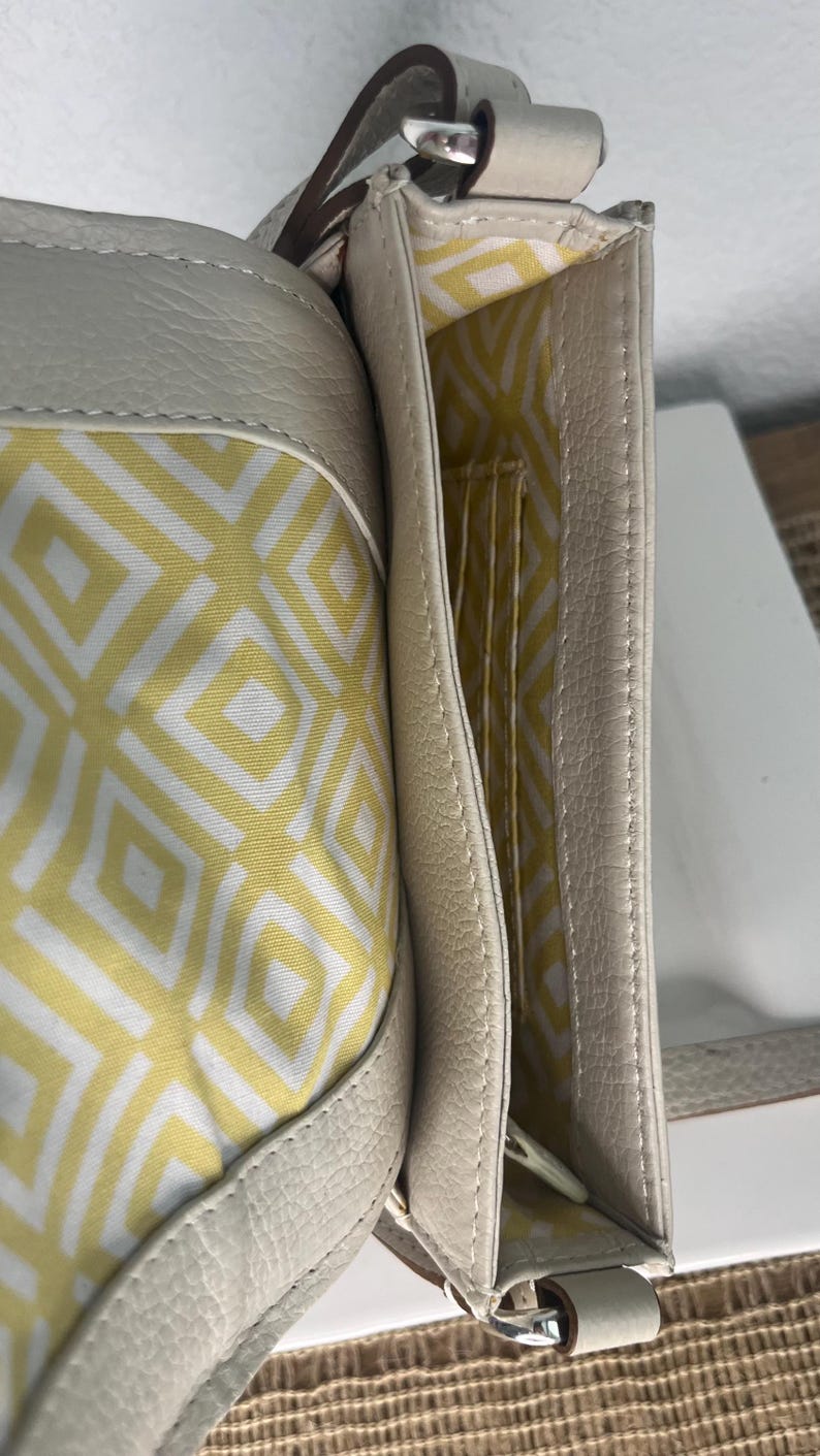 Pode incluir: Bolsa crossbody de couro creme com interior estampado geom&eacute;trico amarelo e branco. A bolsa possui fecho de aba, al&ccedil;a de ombro e v&aacute;rios compartimentos internos.