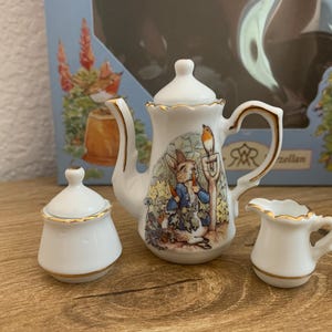 May include: A white porcelain tea set featuring a teapot, sugar bowl, and creamer. The teapot has a detailed illustration of a rabbit in a blue jacket. Gold trim accents the edges of each piece. The set is approximately 4 inches tall.