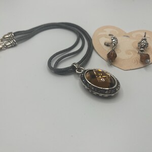 May include: A silver-toned necklace with a large, oval, amber-colored pendant and matching earrings. The pendant and earrings have a decorative silver border. The necklace has a gray cord. The earrings are on a heart-shaped tag.