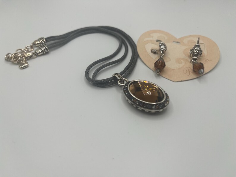 May include: A silver-toned necklace with a large, oval, amber-colored pendant and matching earrings. The pendant and earrings have a decorative silver border. The necklace has a gray cord. The earrings are on a heart-shaped tag.