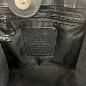 May include: Interior view of a black leather handbag. The bag features a zipper, a magnetic closure, and a label with the Brighton logo and text. The interior is lined with black fabric.