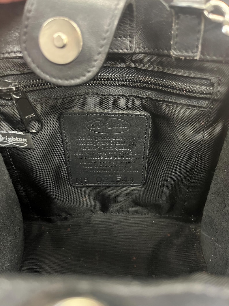 May include: Interior view of a black leather handbag. The bag features a zipper, a magnetic closure, and a label with the Brighton logo and text. The interior is lined with black fabric.