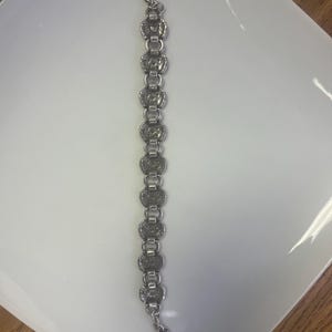 Puede incluir: Pulsera plateada con una serie de eslabones circulares ornamentados. Cada eslab&oacute;n tiene un dise&ntilde;o detallado, conectado por eslabones rectangulares m&aacute;s peque&ntilde;os. La pulsera tiene un cierre de broche y un peque&ntilde;o dije.