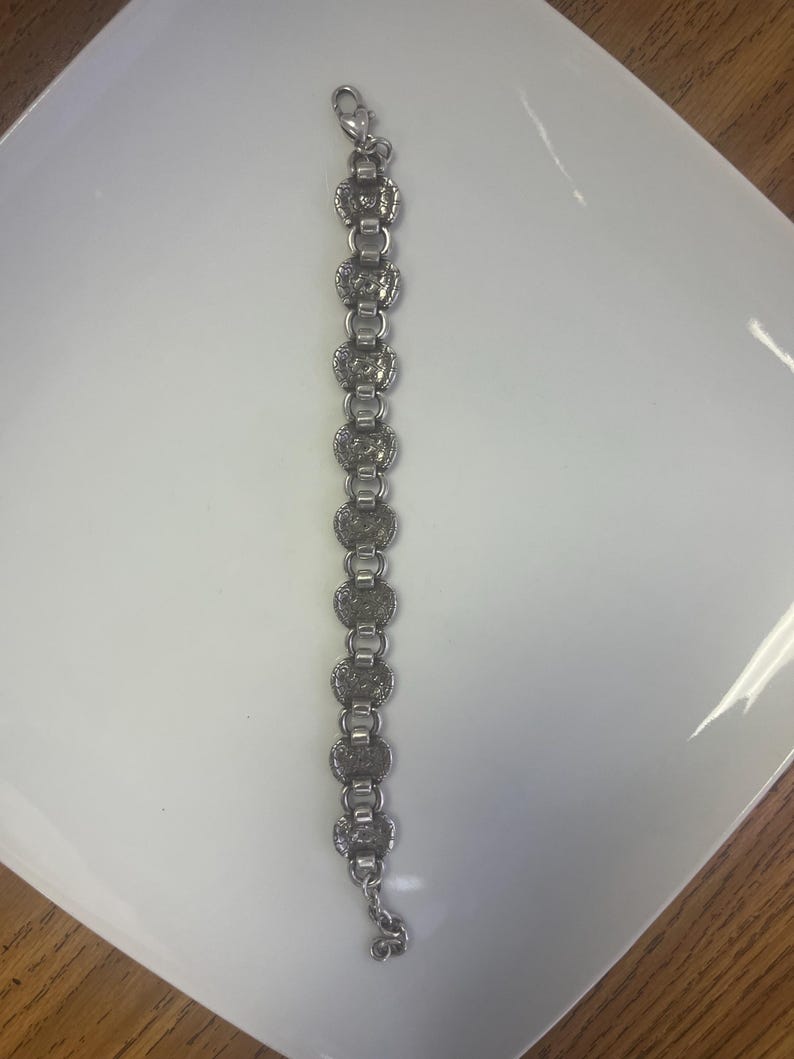 Puede incluir: Pulsera plateada con una serie de eslabones circulares ornamentados. Cada eslab&oacute;n tiene un dise&ntilde;o detallado, conectado por eslabones rectangulares m&aacute;s peque&ntilde;os. La pulsera tiene un cierre de broche y un peque&ntilde;o dije.