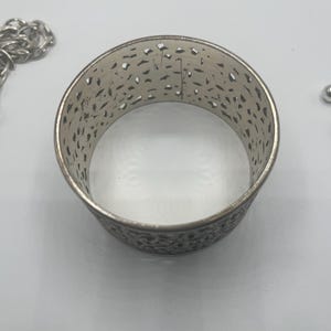 May include: Silver-toned metal cuff bracelet with an openwork design. The bracelet has a wide band with intricate cut-out patterns. A silver chain is visible on the left side of the image, and a small silver bead is on the right.