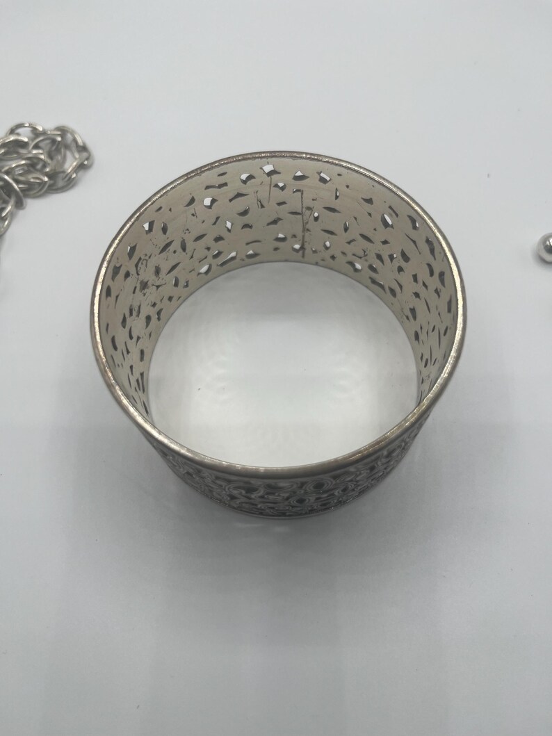 May include: Silver-toned metal cuff bracelet with an openwork design. The bracelet has a wide band with intricate cut-out patterns. A silver chain is visible on the left side of the image, and a small silver bead is on the right.