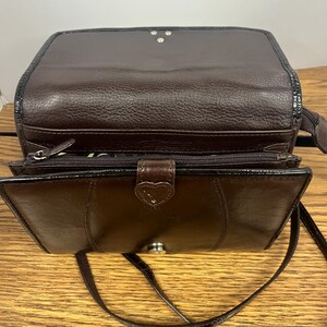 May include: A brown leather crossbody bag with a flap closure and a long, thin strap. The bag has a textured exterior and a heart-shaped clasp. The interior is visible, revealing a zippered compartment.