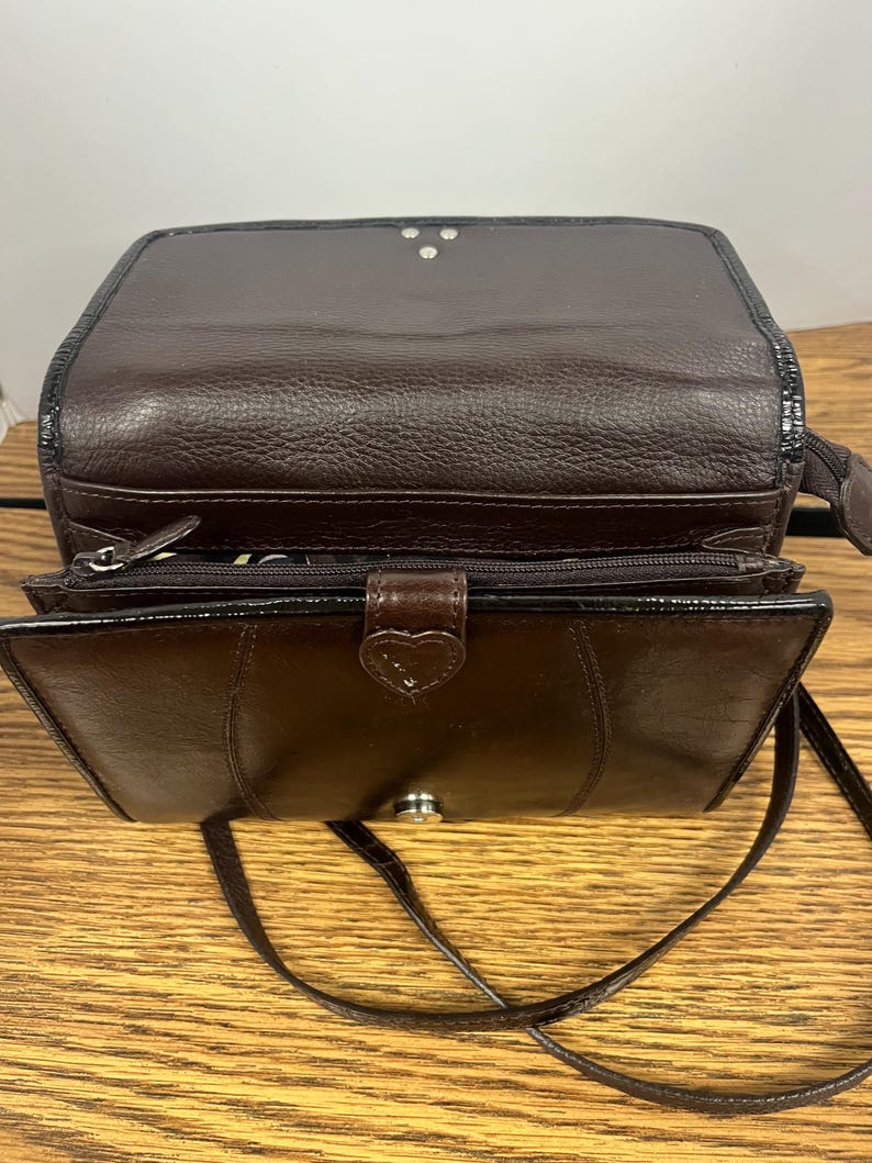May include: A brown leather crossbody bag with a flap closure and a long, thin strap. The bag has a textured exterior and a heart-shaped clasp. The interior is visible, revealing a zippered compartment.