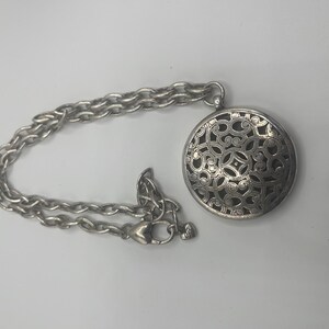 May include: Silver-toned necklace featuring a round pendant with an intricate openwork design. The pendant is connected to a chain with a lobster clasp closure. The necklace is displayed on a white surface.
