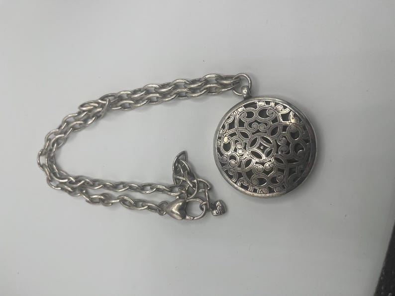 May include: Silver-toned necklace featuring a round pendant with an intricate openwork design. The pendant is connected to a chain with a lobster clasp closure. The necklace is displayed on a white surface.