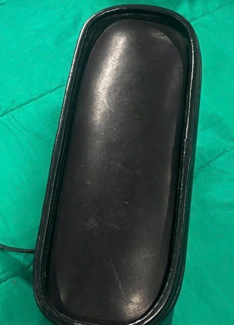 May include: A black, rectangular eyeglasses case with a rounded top. The case is made of a smooth, leather-like material and has a black frame. The case is empty.