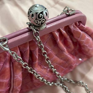 May include: Pink clutch with a silver chain strap. The clutch has a textured, crocodile-like pattern and a decorative silver clasp. The clasp features an ornate, filigree design. The bag is a soft pink color.