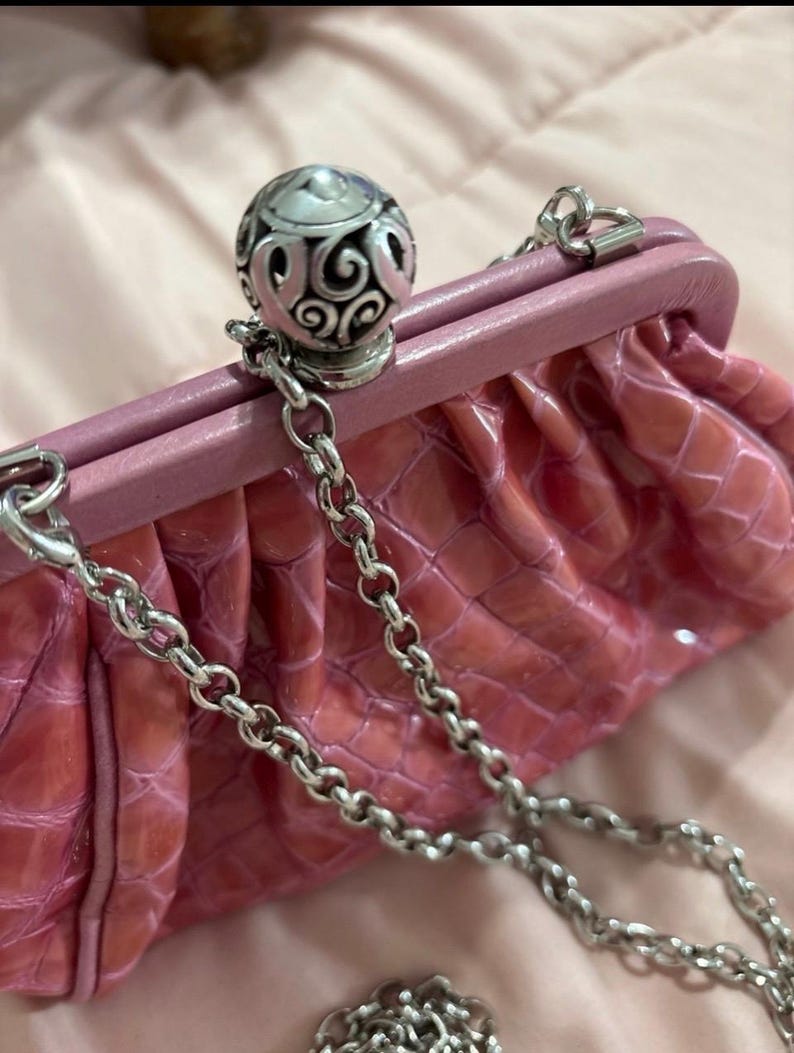 May include: Pink clutch with a silver chain strap. The clutch has a textured, crocodile-like pattern and a decorative silver clasp. The clasp features an ornate, filigree design. The bag is a soft pink color.