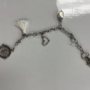 May include: Silver chain accessory with a compass charm, a heart charm, and a white tassel. The chain includes a lobster clasp and an oval bead. This item is likely a keychain or bag charm.