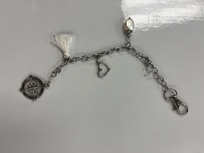 May include: Silver chain accessory with a compass charm, a heart charm, and a white tassel. The chain includes a lobster clasp and an oval bead. This item is likely a keychain or bag charm.