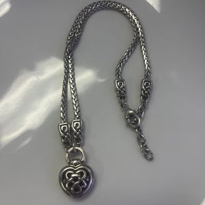 Puede incluir: Collar plateado con dise&ntilde;o de cadena trenzada y colgante en forma de coraz&oacute;n. El colgante tiene un dise&ntilde;o calado. El collar se muestra sobre una superficie blanca.