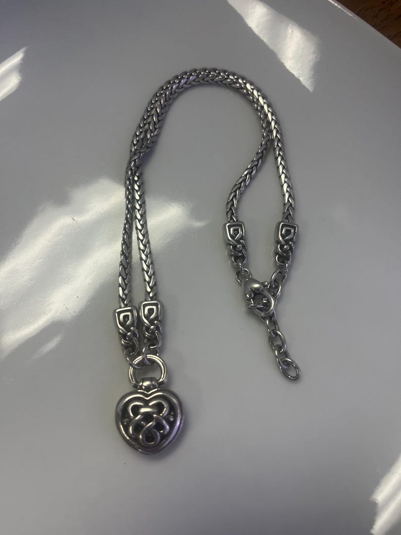 Puede incluir: Collar plateado con dise&ntilde;o de cadena trenzada y colgante en forma de coraz&oacute;n. El colgante tiene un dise&ntilde;o calado. El collar se muestra sobre una superficie blanca.