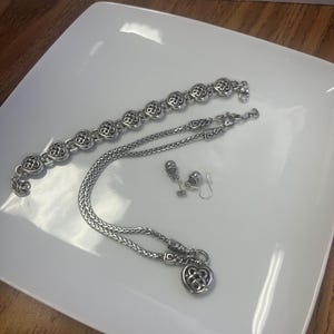Puede incluir: Un conjunto de joyas plateadas que incluye una pulsera, un collar y pendientes. La pulsera y el collar tienen un dise&ntilde;o floral repetido, mientras que el collar tiene un colgante en forma de coraz&oacute;n. Los pendientes son peque&ntilde;os tachuelas. Las joyas se exhiben en un plato blanco.