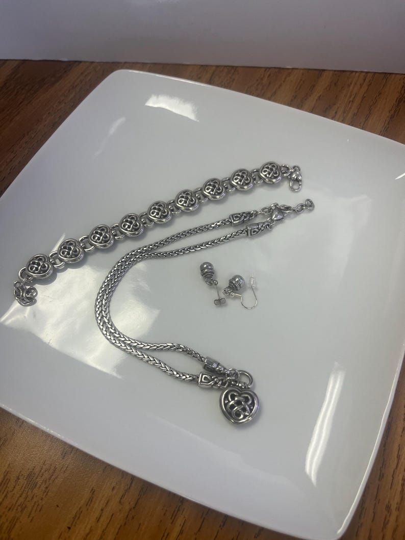 Puede incluir: Un conjunto de joyas plateadas que incluye una pulsera, un collar y pendientes. La pulsera y el collar tienen un dise&ntilde;o floral repetido, mientras que el collar tiene un colgante en forma de coraz&oacute;n. Los pendientes son peque&ntilde;os tachuelas. Las joyas se exhiben en un plato blanco.