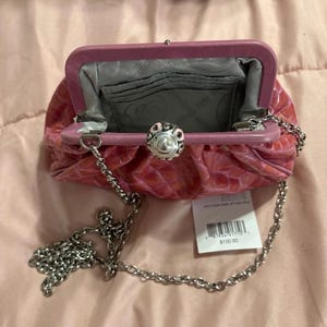 May include: Pink clutch purse with a silver chain strap. The purse has a textured, crocodile-like pattern and a decorative silver clasp. The interior is gray with card slots. A price tag is attached.