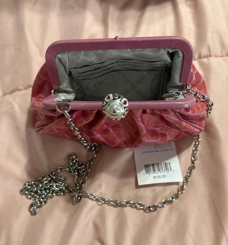 May include: Pink clutch purse with a silver chain strap. The purse has a textured, crocodile-like pattern and a decorative silver clasp. The interior is gray with card slots. A price tag is attached.
