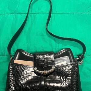 May include: Black handbag with a crocodile-textured exterior and a shoulder strap. The bag features a flap closure with a decorative silver-tone clasp. The interior is visible, with a card and a clear plastic sleeve.