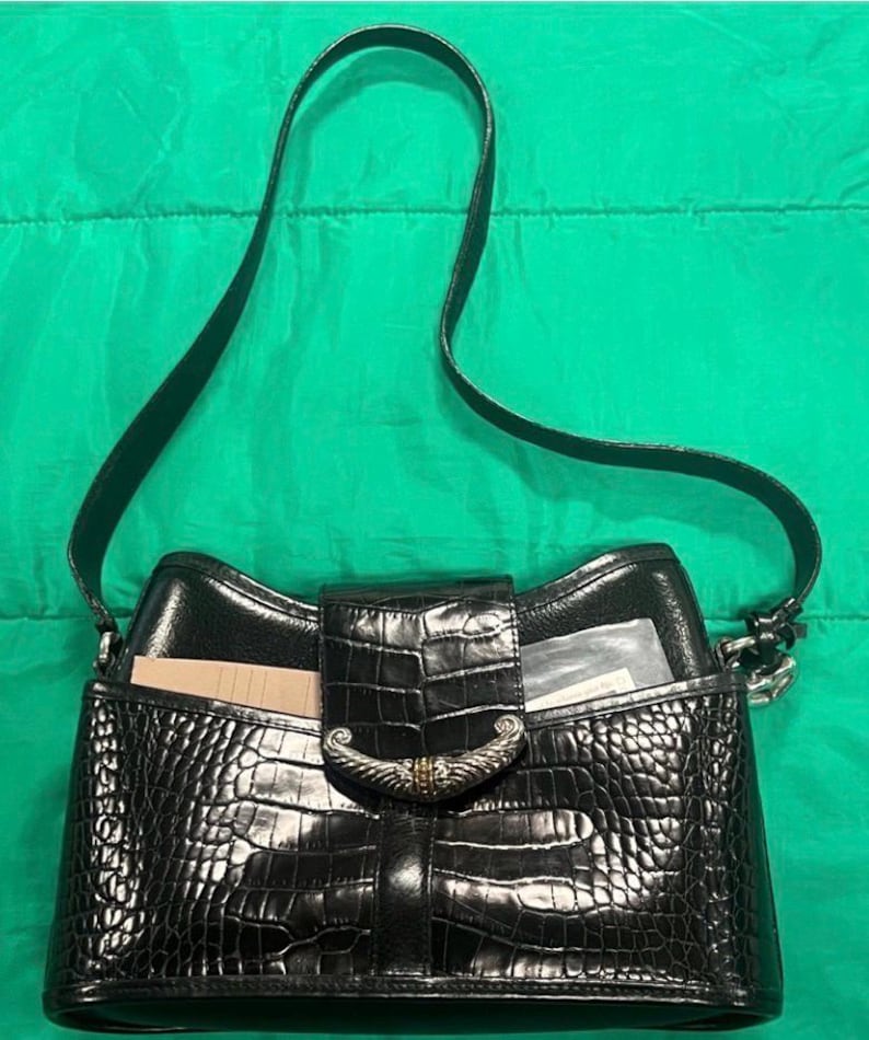 May include: Black handbag with a crocodile-textured exterior and a shoulder strap. The bag features a flap closure with a decorative silver-tone clasp. The interior is visible, with a card and a clear plastic sleeve.