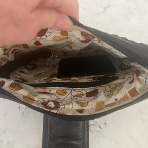 Puede incluir: Bolso de mano de cuero negro con interior estampado. El interior presenta una tela color crema con un dise&ntilde;o floral y de alimentos. El bolso tiene m&uacute;ltiples compartimentos y un cierre de cremallera.