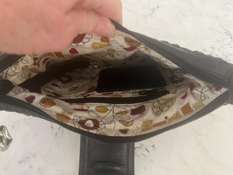 Puede incluir: Bolso de mano de cuero negro con interior estampado. El interior presenta una tela color crema con un dise&ntilde;o floral y de alimentos. El bolso tiene m&uacute;ltiples compartimentos y un cierre de cremallera.