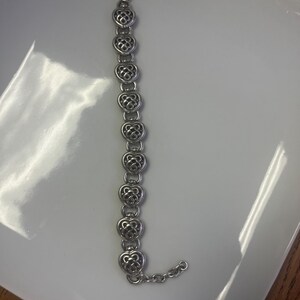 Puede incluir: Una pulsera plateada con eslabones en forma de coraz&oacute;n. Cada coraz&oacute;n presenta un dise&ntilde;o calado intrincado. La pulsera se muestra sobre una superficie blanca, resaltando sus delicados detalles y el cierre de cadena.